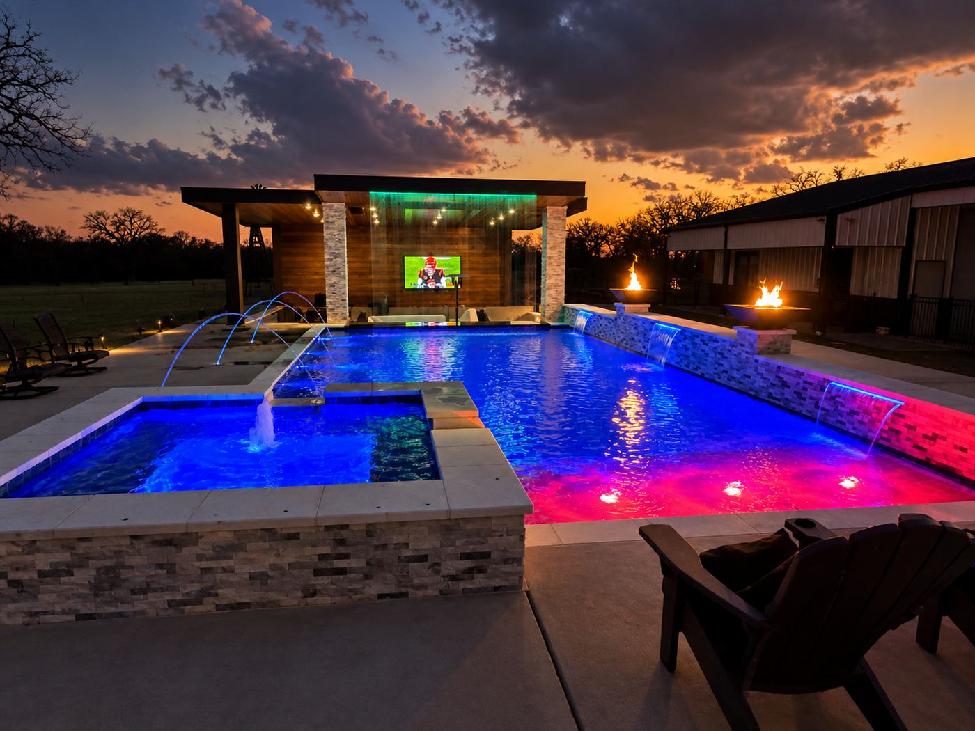 Custom water features at dusk — laminar deck jets, sheer descents spilling from a raised stone wall, a spillover spa, fire bowls, and a covered pavilion with an outdoor TV