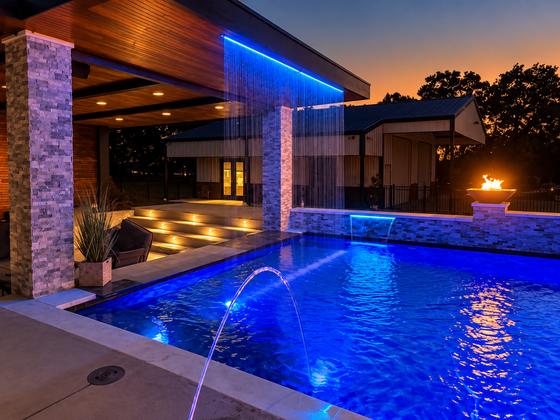 Custom gunite pool at twilight with deck jets, LED lighting and a fire bowl beside a covered patio
