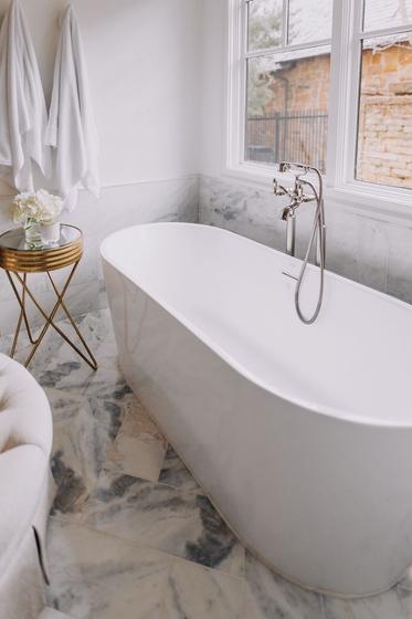Freestanding soaking tub beneath a window in a marble primary bathroom